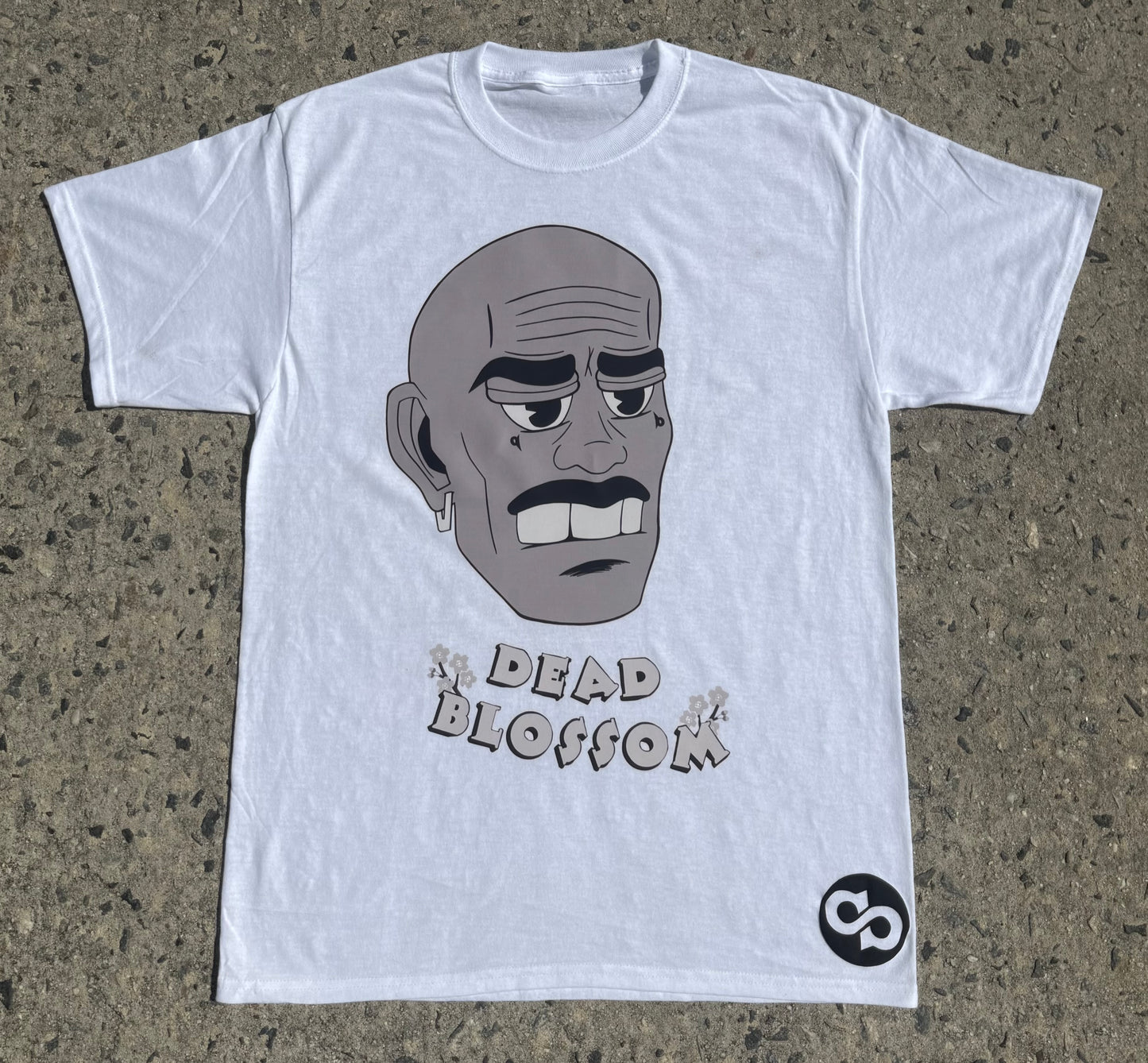 White t-shirt with a graphic of a face and 'The Dead Blossom' text on a concrete surface